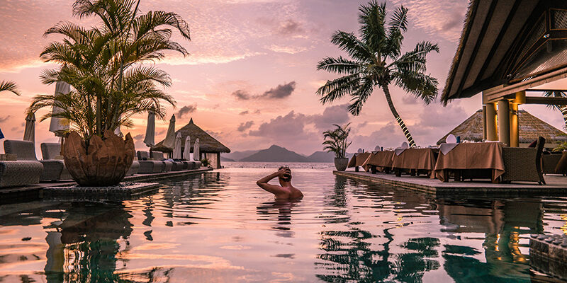 person in swimming pool in luxury resort with palm treas and beach in background